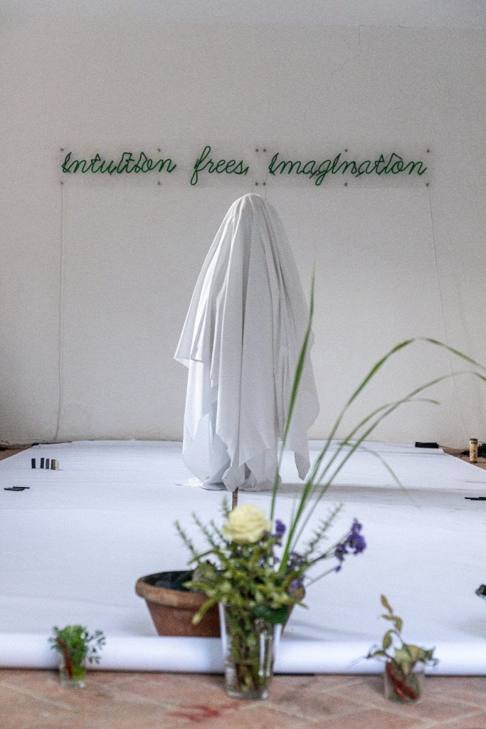 The image shows a person hidden underneath a white blanket, standing on a long and wide sheet of paper. On the wall in the background it says: "intuition frees imagination".
