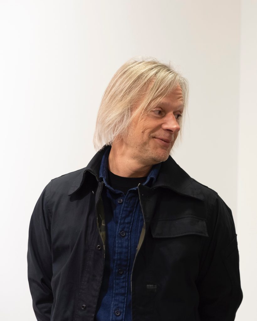 A middle-aged man with shoulder-length white hair smiles gently as he turns his gaze to the side.
